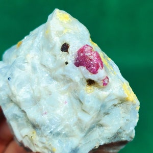 May include: A white, gray, and yellow rock with a bright pink ruby crystal embedded in it.
