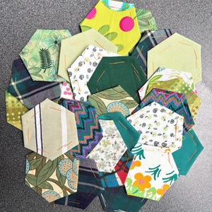 Op de afbeelding: Een verzameling kleurrijke, zeshoekige stoffen stukken in verschillende patronen en tinten groen, geel en blauw. Sommige stukken hebben bloemen-, ruit- en geometrische ontwerpen, terwijl andere effen kleuren hebben. De stukken zijn in een cirkelvorm gerangschikt.