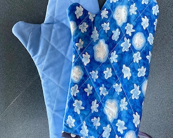 May include: Two quilted oven mitts. One is solid light blue with a diamond pattern. The other is a vibrant blue with a floral pattern in white and light blue. Both mitts have a loop for hanging and a dark blue trim.