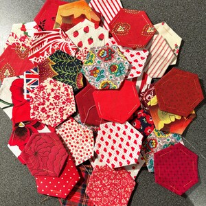Op de afbeelding: Een verzameling rode en witte stoffen zeshoeken met verschillende patronen, waaronder strepen, bloemen, aardbeien en stippen. De patchwork stukken zijn in een cirkelvorm gerangschikt en tonen een verscheidenheid aan texturen en ontwerpen.