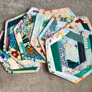 May include: A collection of handmade, hexagonal fabric coasters. Each coaster features a patchwork design with various patterns, including floral, animal prints, and geometric shapes. The coasters have a light-colored border and are arranged on a neutral surface.