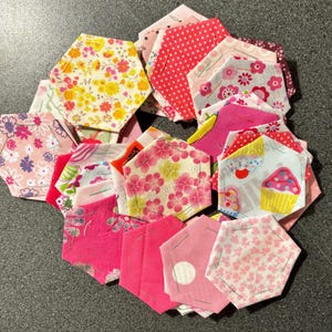 May include: A circular arrangement of hexagonal fabric patches in various shades of pink, red, and white. The patches display floral and polka dot patterns, with some featuring cupcake designs. Each piece is made of fabric.