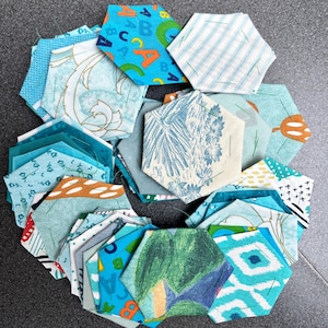 May include: A circular arrangement of hexagonal fabric pieces in various patterns and colours. The fabrics display floral, geometric, and letter designs in shades of blue, green, and orange. These pieces are likely for quilting or crafting projects.