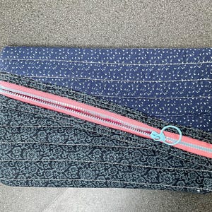Puede incluir: Una bolsa de tela azul y negra con cremallera rosa y tirador de cremallera azul. La bolsa tiene un patrón floral en un lado y un pequeño patrón de puntos blancos en el otro.