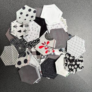 Op de afbeelding: Een verzameling van diverse stoffen zeshoeken in verschillende patronen en kleuren. Ontwerpen omvatten stippen, bloemen en geometrische vormen in zwart, wit, grijs en rood. De zeshoeken zijn gerangschikt op een grijze ondergrond.