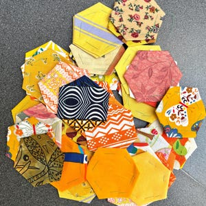 Op de afbeelding: Een verzameling kleurrijke, zeshoekige stofstukken in verschillende patronen en tinten geel, oranje en bloemenprints. Sommige stukken hebben geometrische ontwerpen, andere hebben vlinders en bladeren.