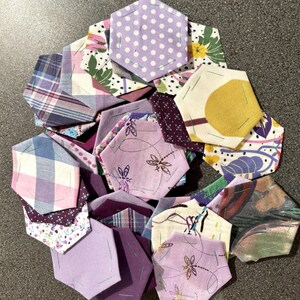 Op de afbeelding: Een verzameling zeshoekige stoffen stukken in verschillende patronen en tinten paars, lavendel en wit. Sommige stukken hebben bloemen-, stippen- en ruitpatronen. De stukken zijn in een cirkelvorm gerangschikt.