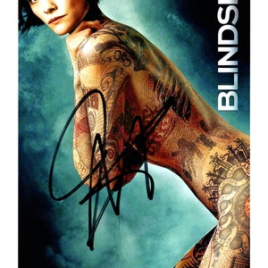 Può includere: Una donna con i capelli corti e scuri è ricoperta di intricati tatuaggi. Sta posando per un servizio fotografico. Lo sfondo è un gradiente blu e verde. Il testo "BLINDSPOT" è scritto verticalmente in lettere bianche sul lato destro dell'immagine.