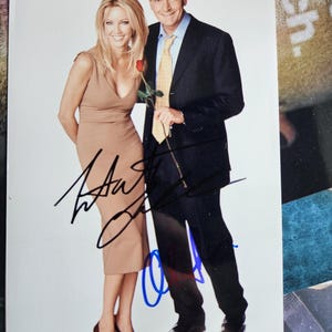 Puede incluir: Fotografía firmada de un hombre y una mujer. La mujer viste un vestido color beige, el hombre un traje negro con corbata amarilla, sosteniendo una rosa roja. La foto está firmada con tinta negra y azul.