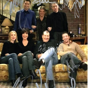 Könnte beinhalten: Ein Gruppenfoto von sieben Personen, vermutlich einer Besetzung oder Crew, die auf einem blumigen Sofa posieren. Das Bild ist mit mehreren Autogrammen in weißer Tinte signiert. Die Personen sind in Freizeitkleidung gekleidet, und der Hintergrund umfasst eine Lampe und eine Backsteinmauer.