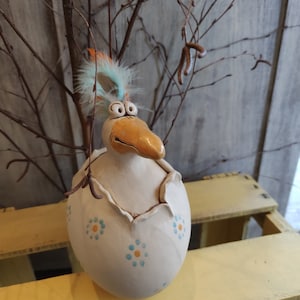 Könnte beinhalten: Eine skurrile Keramikfigur eines Vogels, der aus einer zerbrochenen Eierschale schlüpft. Der Vogel hat einen großen orangen Schnabel, große Augen und einen Büschel blauer und orangefarbener Federn. Die Eierschale ist weiß mit blauen und gelben Blumenmustern.