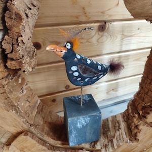 Può includere: Una stravagante scultura di uccello in legno dipinta a mano. L'uccello presenta un corpo nero con pois bianchi, un becco arancione e una coda soffice. È montato su un blocco grigio e posto su uno sfondo di legno, incorniciato da una fetta di legno naturale.