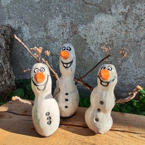 Könnte beinhalten: Drei skurrile Schneemannfiguren mit Zweigarmen und getrockneten Blumendetails. Jeder Schneemann hat eine Karottennase, einen lächelnden Mund und Knopffunktionen. Die Schneemänner werden auf einer Holzoberfläche ausgestellt.