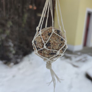 Könnte beinhalten: Eine kugelförmige Vogelfutterkugel aus Samen, Getreide und getrockneten Beeren, die in einem weißen, geknoteten Makramee-Netz aus Schnur steckt. Das Netz hat rautenförmige Öffnungen und ist am unteren Ende mit einem Knoten versehen.
