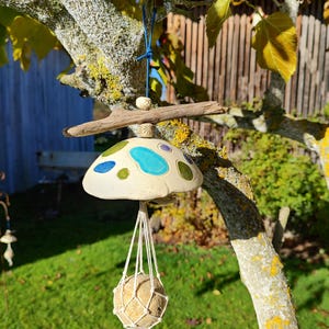 Op de afbeelding: Een handgemaakte keramische paddenstoel vogelvoeder met een beige kap, versierd met blauwe en groene details. Een macrame net houdt een ronde, getextureerde vogelvoerbal vast. De voederbak hangt aan een tak, wat een charmant tuinornament creëert.