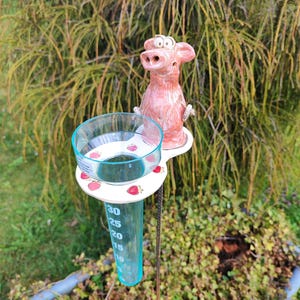 Könnte beinhalten: Eine skurrile rosa Keramik-Kuhfigur sitzt auf einer weißen Plattform mit einem transparenten Regenmesser. Die Anzeige hat blaue Markierungen zur Messung des Niederschlags in Millimetern. Die Plattform ist mit roten Erdbeermotiven verziert. Der Regenmesser ist auf einer Metallstange montiert.