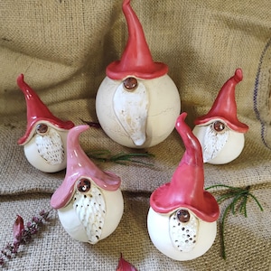 Può includere: Cinque figurine di gnomi in ceramica. Ogni gnomo ha un corpo rotondo bianco sporco, un cappello a punta rosso o rosa e una barba bianca. I cappelli hanno una piccola gemma marrone. Gli gnomi sono disposti su una superficie beige strutturata.