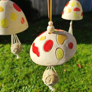 Pode incluir: Sinos de vento em forma de cogumelo de cerâmica com detalhes em vermelho e amarelo. Cada sino tem uma tampa bege com manchas coloridas e uma esfera suspensa. Os sinos são suspensos por cordas amarelas.