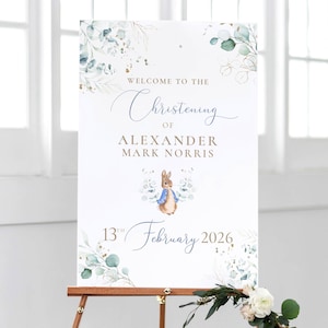 May include: A white sign on a wooden easel, announcing "Welcome to the Christening of Alexander Mark Norris 13th February 2026." The sign is decorated with a Peter Rabbit illustration and floral elements.