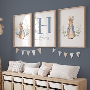 May include: Three framed prints with a blue and white watercolour design featuring a bunny rabbit and the letter H with the name Harry. The prints are hanging on a blue wall above a wooden bench with cushions.