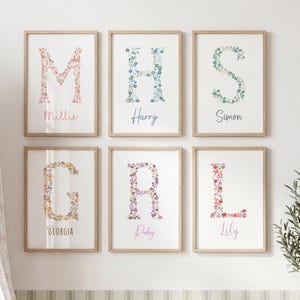 May include: Six framed wall art prints featuring floral-patterned letters M, H, S, G, R, and L. Each print includes a name written below the letter: Millie, Harry, Simon, Georgia, Ruby, and Lily. The frames are light wood, and the prints are white.