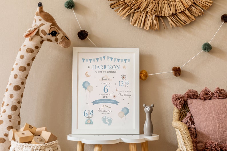 May include: A white framed print with blue and white illustrations of a moon, stars, balloons, and a clock. The print includes the text "HARRISON George Dunne Born on Monday 6th February 2023 at 12:43 am Proud Parents Alice & George 6.8 lbs Harrison Park Hospital".