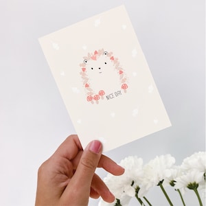 Könnte beinhalten: Eine weiße Karte mit einer Illustration eines rosa-braunen Igels, umgeben von Blättern und Pilzen. Der Text "NICE DAY" ist unter dem Igel gedruckt.