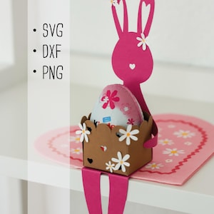 Könnte beinhalten: Ein rosa Osterhase aus Papier, der eine braune Schachtel mit gewelltem Rand hält, verziert mit weißen Papierblumen. Der Hase sitzt auf einer herzförmigen rosa Matte mit Gänseblümchenmustern. Der Text: SVG, DXF, PNG.