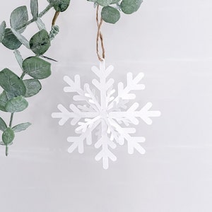 Op de afbeelding: Witte papieren sneeuwvlok ornament hangend aan een touwtje. De sneeuwvlok is gedetailleerd en ingewikkeld met een 3D-effect.