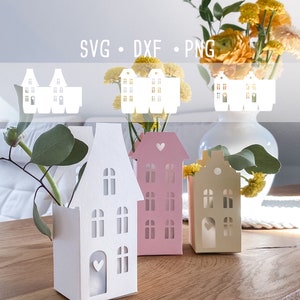 Può includere: Tre vasi di carta a forma di casa in bianco, rosa e giallo con finestre e cuori ritagliati. I vasi sono pieni di fiori e verde. Il testo "SVG - DXF - PNG" è visibile nella parte superiore dell'immagine.