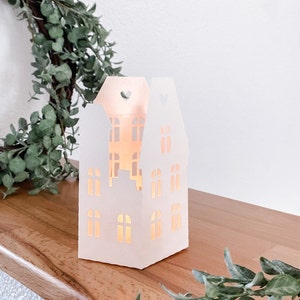 May include: White paper house-shaped candle holder with cut-out windows and a heart at the top. The candle holder is lit and casting a warm glow.