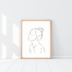 May include: A black and white line drawing of a person's face with their hair pulled back in a bun. The drawing is in a simple, minimalist style and is framed in a light wood frame.