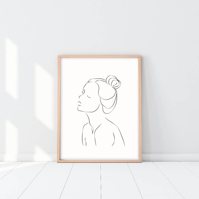 May include: A black and white line drawing of a person's face with their hair pulled back in a bun. The drawing is in a simple, minimalist style and is framed in a light wood frame.