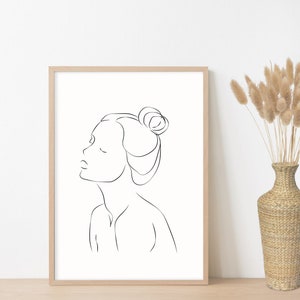 May include: A black and white line drawing of a person's face with their hair pulled back in a bun. The drawing is in a simple, minimalist style and is framed in a light wood frame.