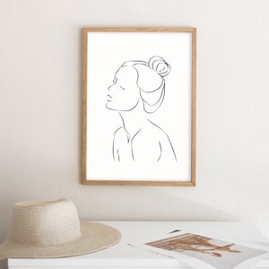 May include: A black and white line drawing of a woman with her hair pulled back in a bun. The drawing is framed in a light wood frame and is hanging on a white wall.