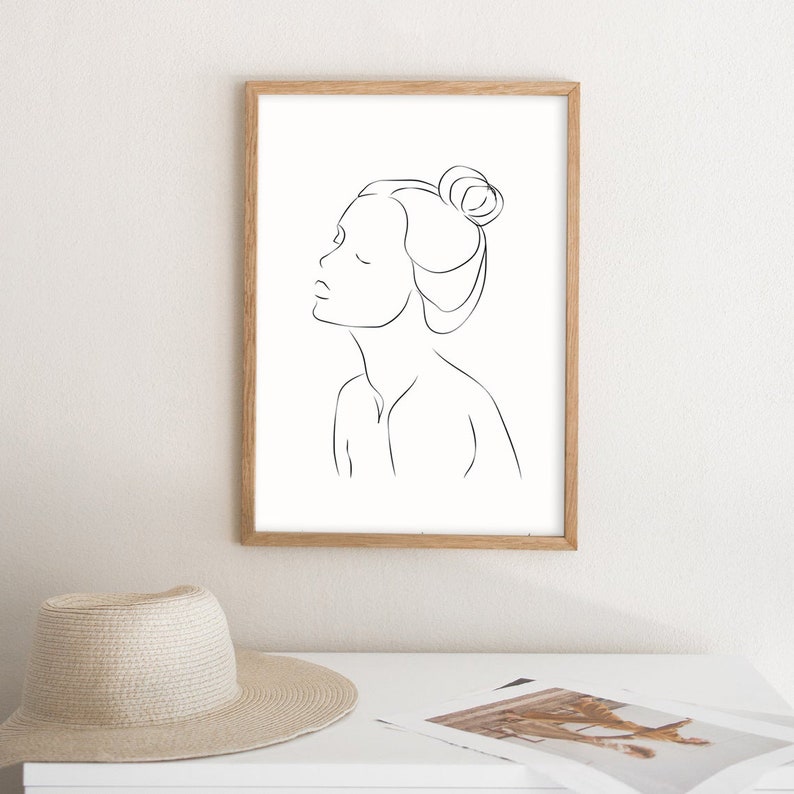 May include: A black and white line drawing of a woman with her hair pulled back in a bun. The drawing is framed in a light wood frame and is hanging on a white wall.