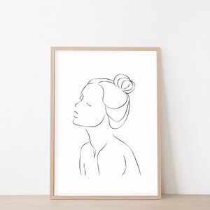 May include: Line art drawing of a person with their hair pulled back in a bun. The drawing is in black and white and is framed in a light wood frame.