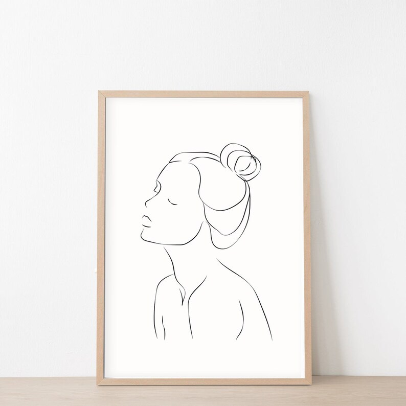 May include: Line art drawing of a person with their hair pulled back in a bun. The drawing is in black and white and is framed in a light wood frame.