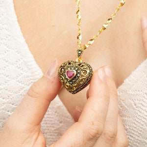 Può includere: Una collana d'oro con un ciondolo a forma di cuore. Il ciondolo è decorato con intricate filigrani e una pietra preziosa rossa al centro. La collana è indossata da una persona con un maglione bianco.