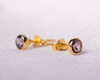 May include: A pair of gold stud earrings with round, light purple gemstones. The earrings are set in a gold bezel setting and have a post and butterfly backing. The earrings are displayed on a white, textured surface.
