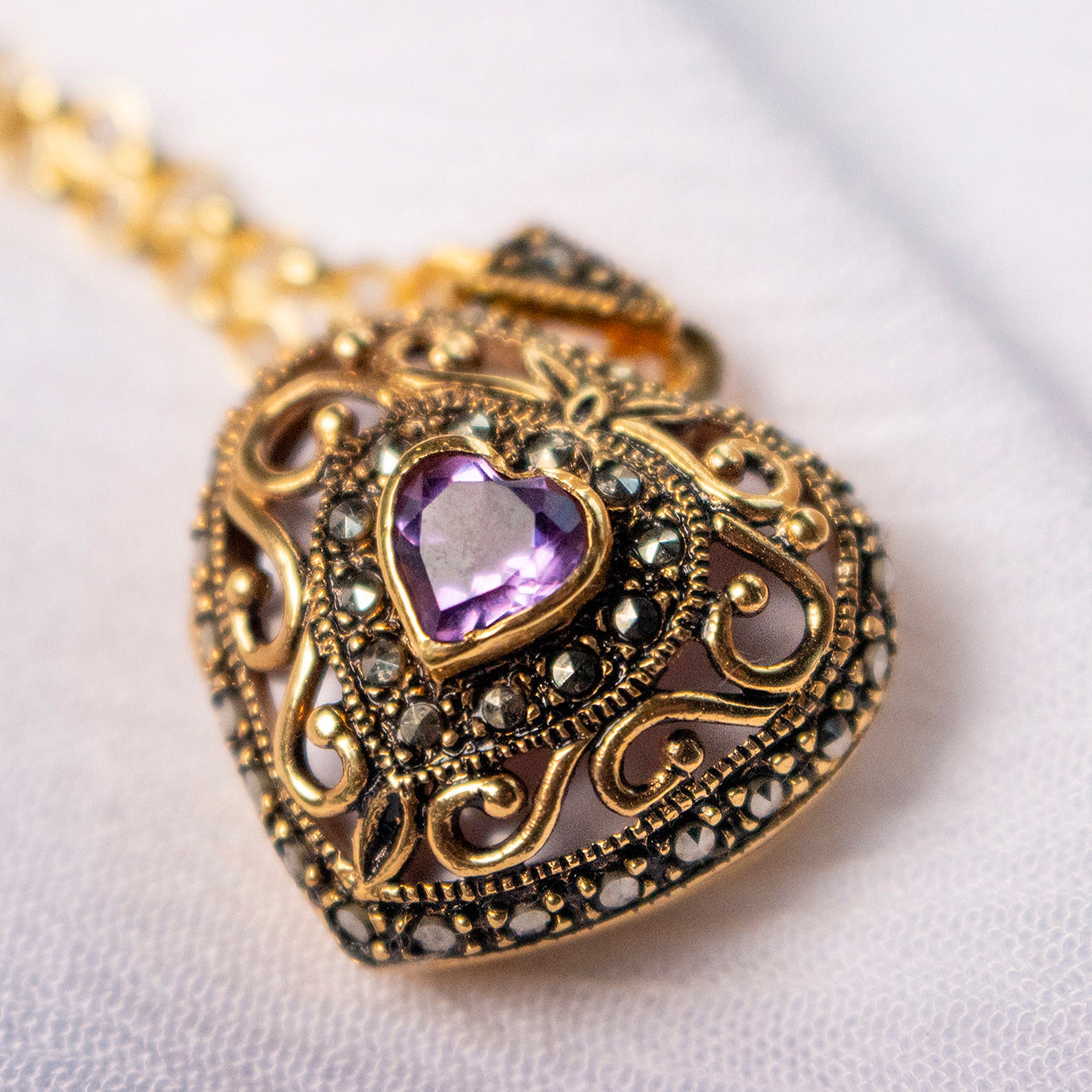 18k gold amethyst heart locket with ornate vintage filigree and marcasite accents on white fabric background.