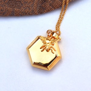 May include: A gold-coloured necklace featuring a hexagonal pendant with a raised bee design. The pendant hangs from a delicate gold chain. The background is white with a brown fabric in the upper left corner.