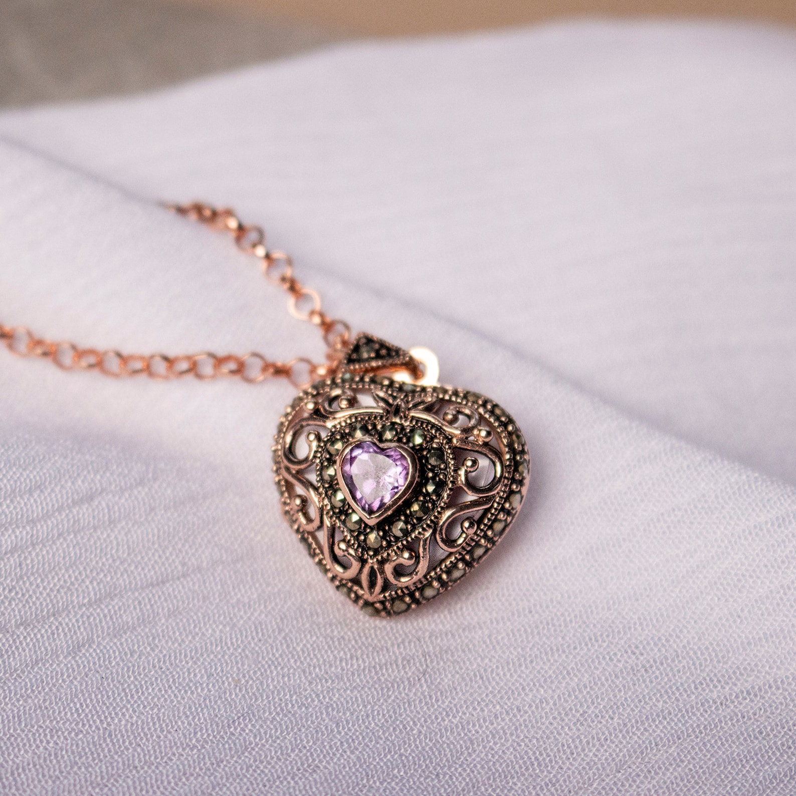 Rose gold amethyst heart locket with filigree detailing and marcasite accents on soft white fabric background.