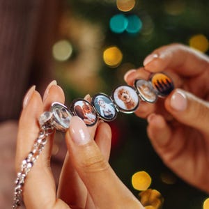 Op de afbeelding: Een zilveren ketting met meerdere bedels, elk met een andere foto. De bedels zijn rond en hebben een zilveren frame. De ketting wordt vastgehouden door iemands handen.