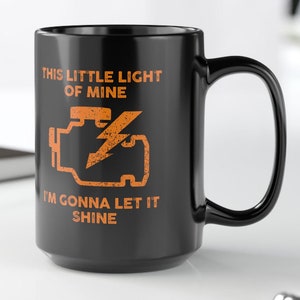 Może przedstawiać: Czarny kubek do kawy z białym nadrukiem, na którym widnieje napis "This little light of mine I'm gonna let it shine" oraz stylizowana pomarańczowa ikona kontrolki silnika.