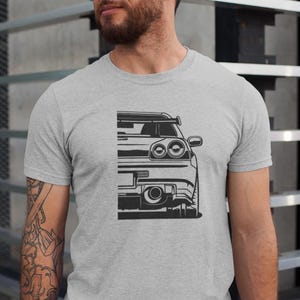 May include: A grey t-shirt with a black and white graphic of a sports car's rear. The design shows the taillights, spoiler, and exhaust. The t-shirt has a crew neck.