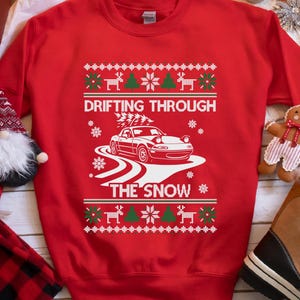 May include: Red sweatshirt featuring a white graphic of a car drifting on a snowy road, with the text "DRIFTING THROUGH THE SNOW". The design includes snowflakes, reindeer, and Christmas trees. A festive Christmas jumper.