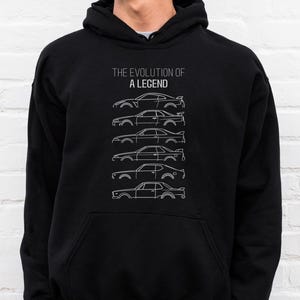 Puede incluir: Sudadera con capucha negra con un gráfico blanco que muestra la evolución de un coche y el texto "THE EVOLUTION OF A LEGEND". El diseño muestra siluetas de coches, ilustrando la evolución del diseño.