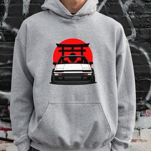 以下が含まれることがあります： 白い車と赤い太陽のグラフィックが描かれた灰色のパーカー。車は日本の門の前にある。グラフィックはパーカーの前面にある。