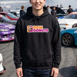 以下が含まれることがあります： オレンジ、白、ピンクで「DOIN DONUTS」の文字が入った黒いフード付きスウェットシャツ。スウェットシャツにはフロントポケットとドローストリングフードが付いています。画像内の人物は笑顔で、複数の車の前に立っています。